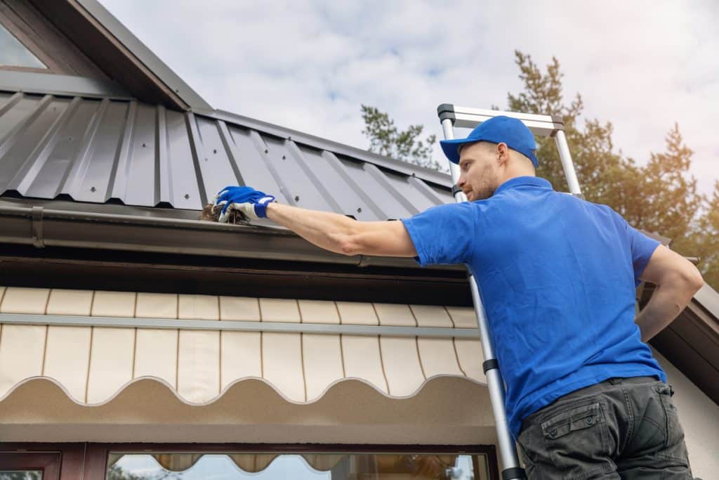 A worker in blue clothing and gloves cleans debris from a roof gutter while standing on a ladder beside a house.